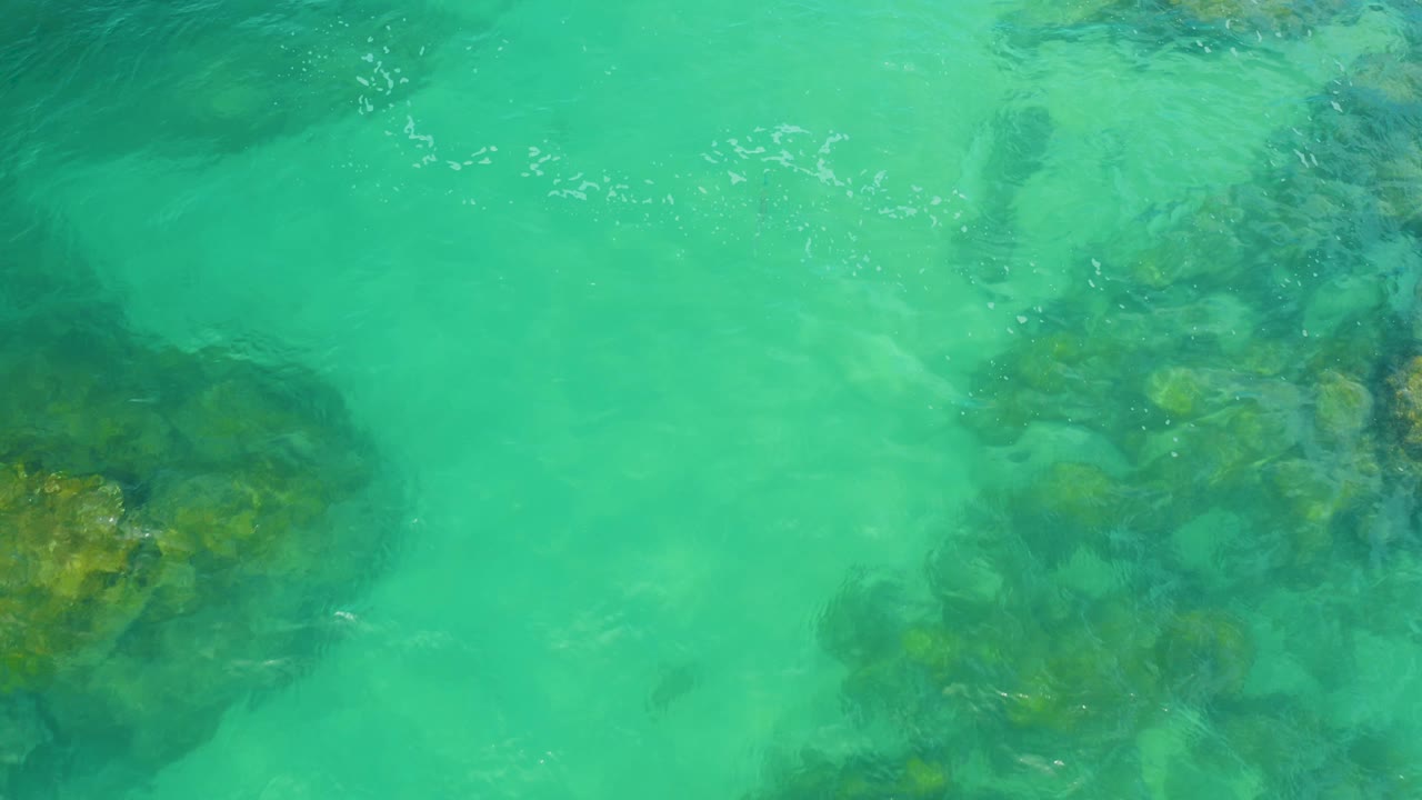 An overhead frame from Laguna Beach, Baja California Sur, shows pale ripples across turquoise water and clustered rocks below, with no visible people.