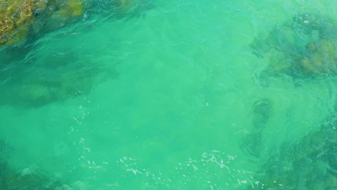 Turquoise coastal water at Laguna Beach, Baja California Sur shows submerged rocks near the upper edge and faint foam lines, with no visible people.