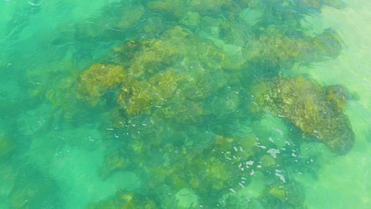 A top-down frame shows pale sand and submerged rock shapes beneath turquoise water in Laguna Beach, Baja California Sur, with no visible people.
