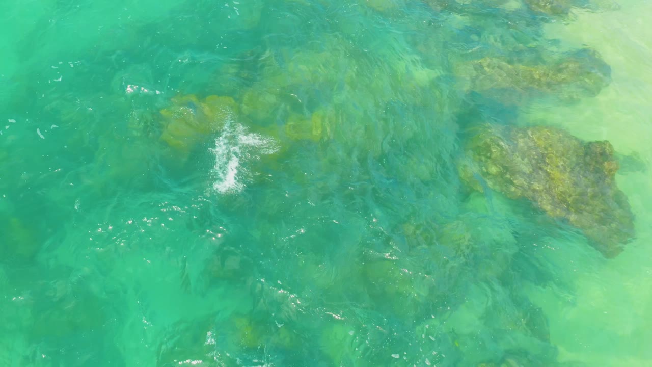 Clear green-blue water covers reef formations at Laguna Beach, Baja California Sur, with bright surface highlights near the left center and no visible people.