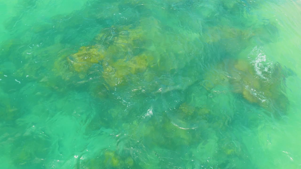 A large submerged reef rock sits under clear green water at Laguna Beach, Baja California Sur, with a small white ripple to the right and no visible people.