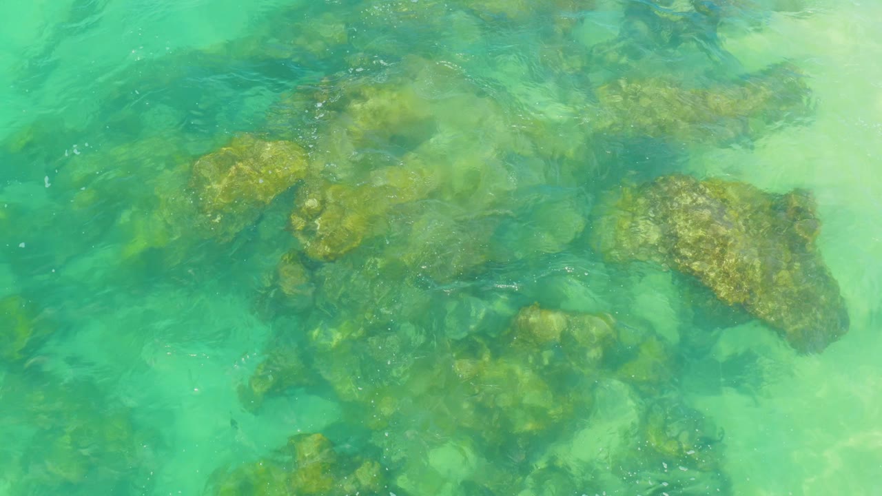 Aerial-style frame of brown rock clusters under clear coastal water in Laguna Beach, Baja California Sur, with pale sand showing through and no visible people.