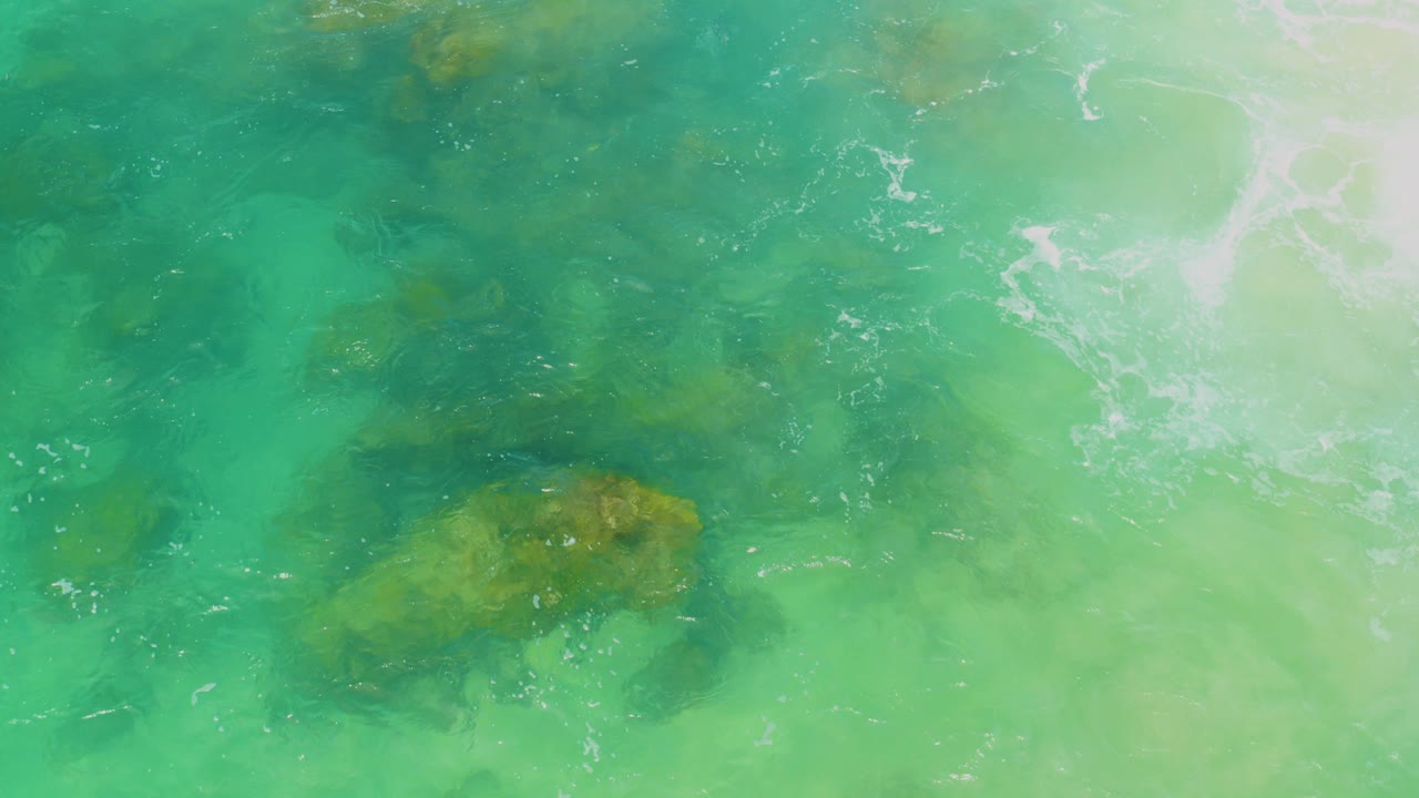 Top-down view of turquoise water over submerged brown rock shapes at Laguna Beach, Baja California Sur, with pale surface glare and no visible people.