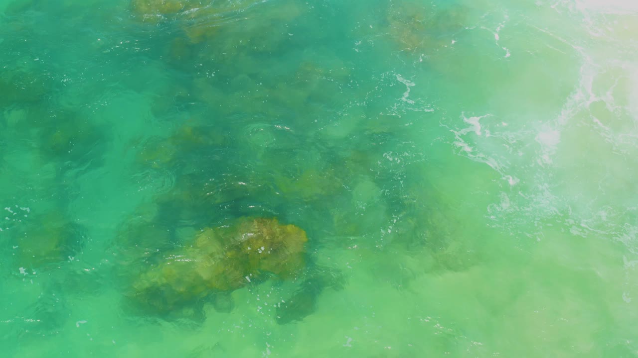 Bright shallow coastal water at Laguna Beach, Baja California Sur, with a brown submerged rock patch below center and soft white turbulence at the upper right, no visible people.