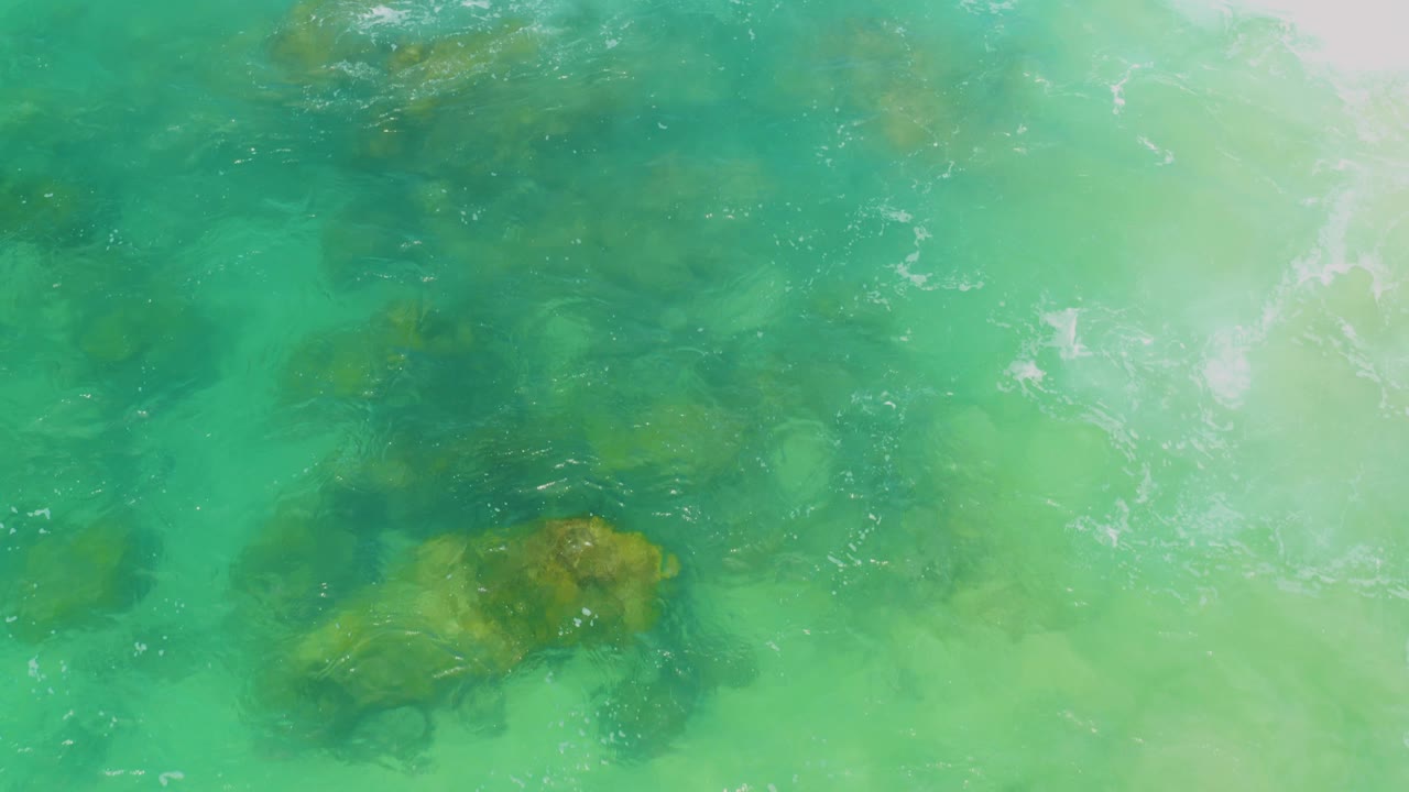 Aquamarine water at Laguna Beach, Baja California Sur, revealing multiple darker submerged patches and one brown rock shape near the lower right, with no visible people.