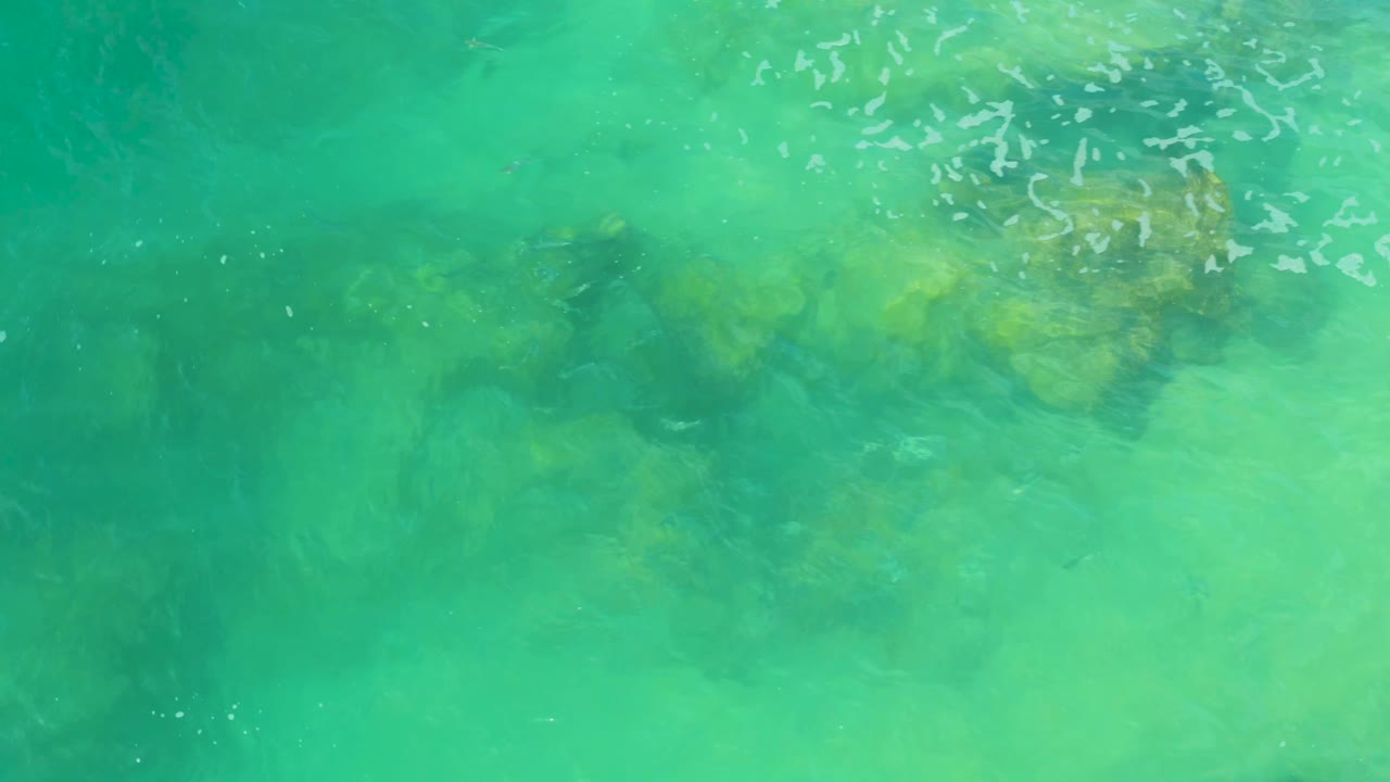 Overhead view of turquoise water at Laguna Beach, Baja California Sur, with dark submerged rocks near the top of frame and no visible people.