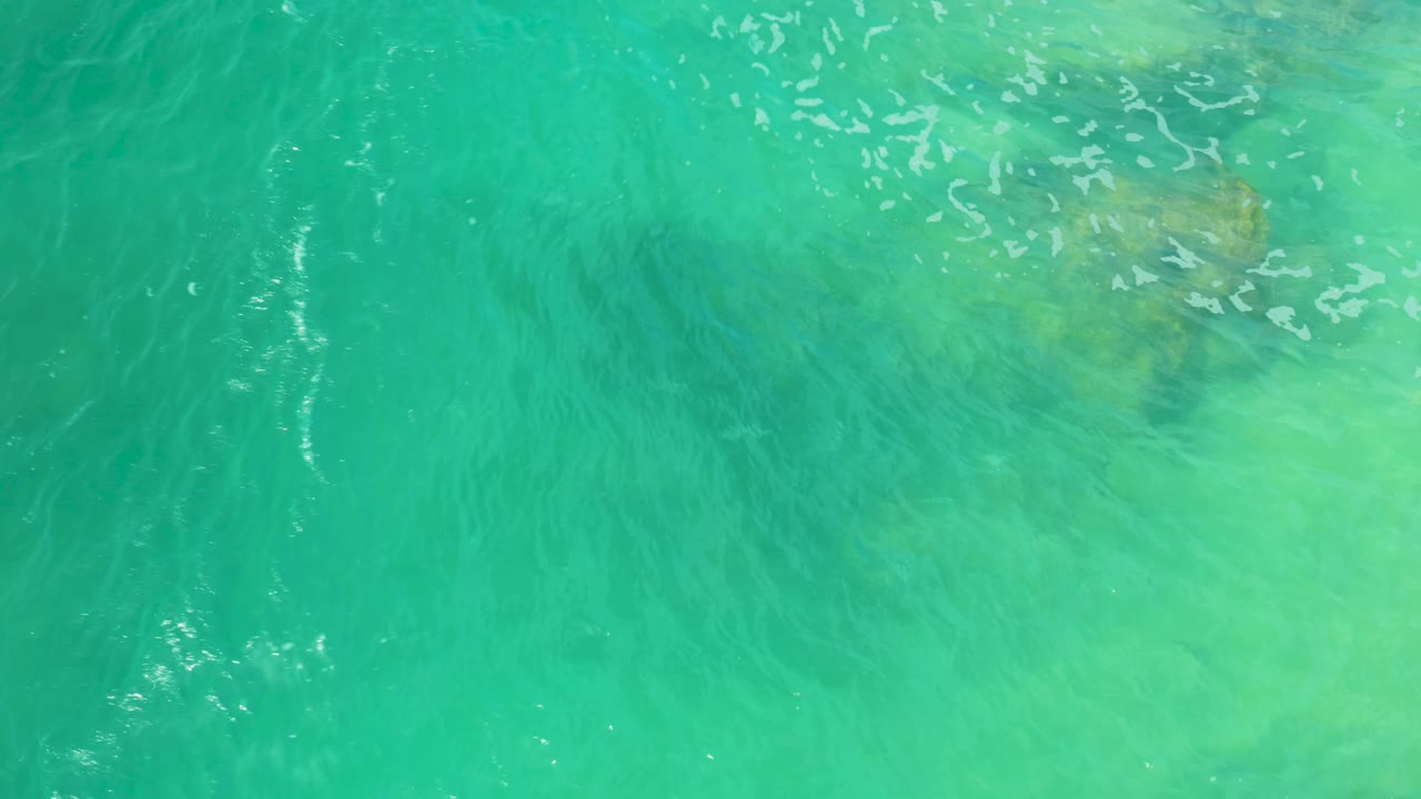 An overhead Laguna Beach water view shows bright ripple marks across turquoise water and muted underwater rocks, with no visible people.