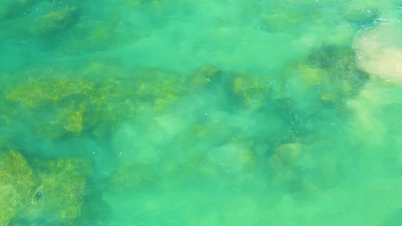 Overhead view of turquoise water at Laguna Beach, Baja California Sur, with submerged rocks, pale sand patches, and no visible people.