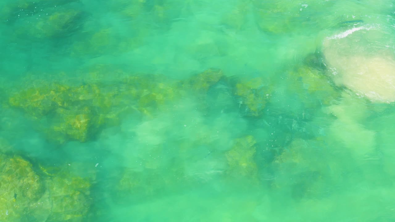 Top-down view of clear green water in Laguna Beach, Baja California Sur, with rocks below and a pale sandy patch near the upper right, no visible people.