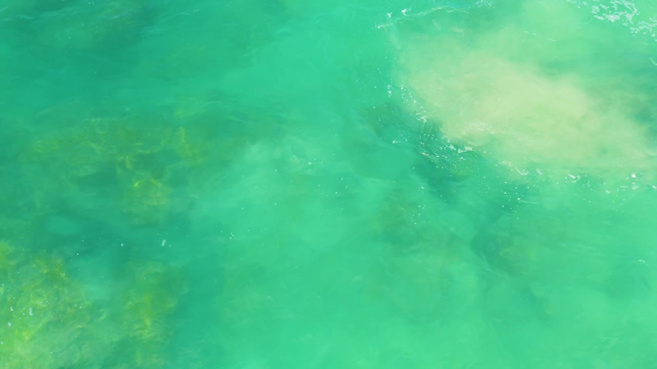 Turquoise coastal water in Laguna Beach, Baja California Sur, with pale foam near the upper right and dim rock forms visible through the water, no visible people.
