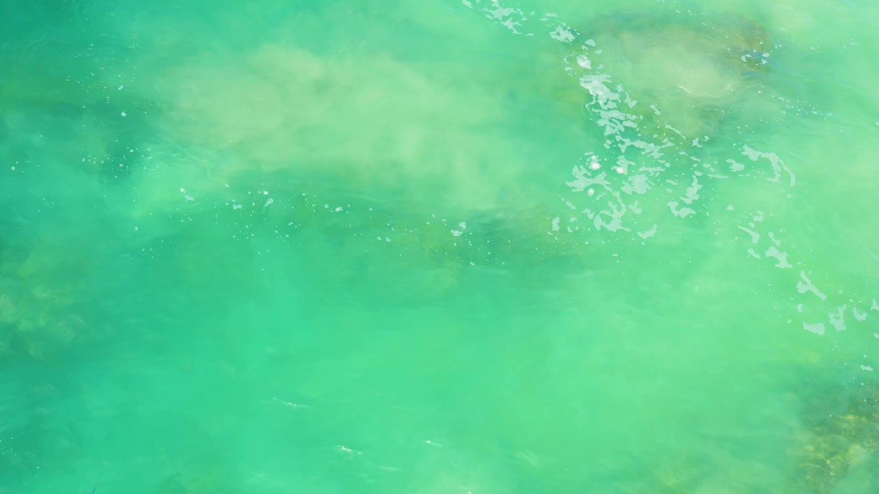 Cloudy green water drifts across darker submerged rock shapes and sandy patches in Laguna Beach, Baja California Sur, with no visible people.