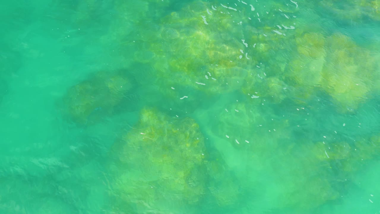 Top-down aerial-looking frame of Laguna Beach water with darker submerged rocks, yellow-green shallows, bright foam flecks near the top right, and no visible people.