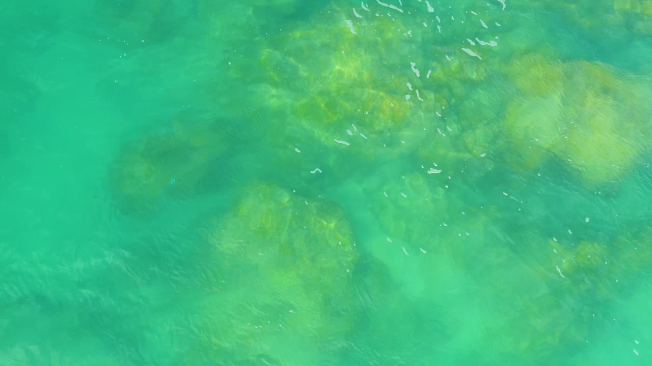 Overhead frame from Laguna Beach, Baja California Sur, showing turquoise water, soft-edged submerged rock shapes, scattered white foam, and no visible people.
