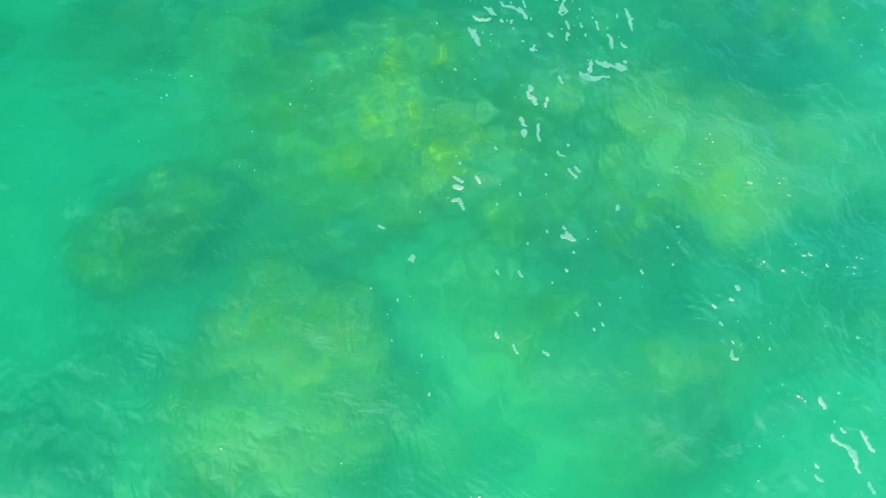 Green sea water in Laguna Beach, Baja California Sur, shows small wavelets, bright white glints, and faint darker shapes below the surface, no visible people.