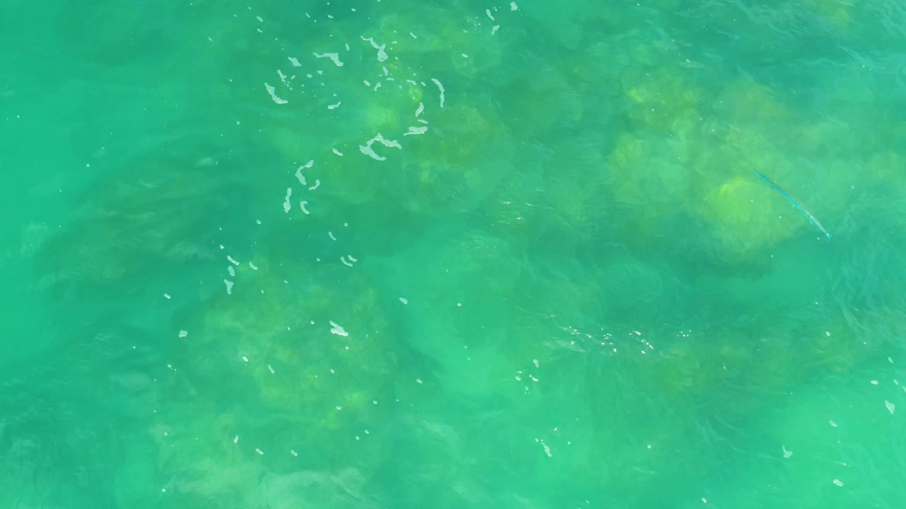 A second overhead frame shows green-blue seawater with small white ripple streaks and muted submerged forms below the surface at Laguna Beach, Baja California Sur, with no visible people.