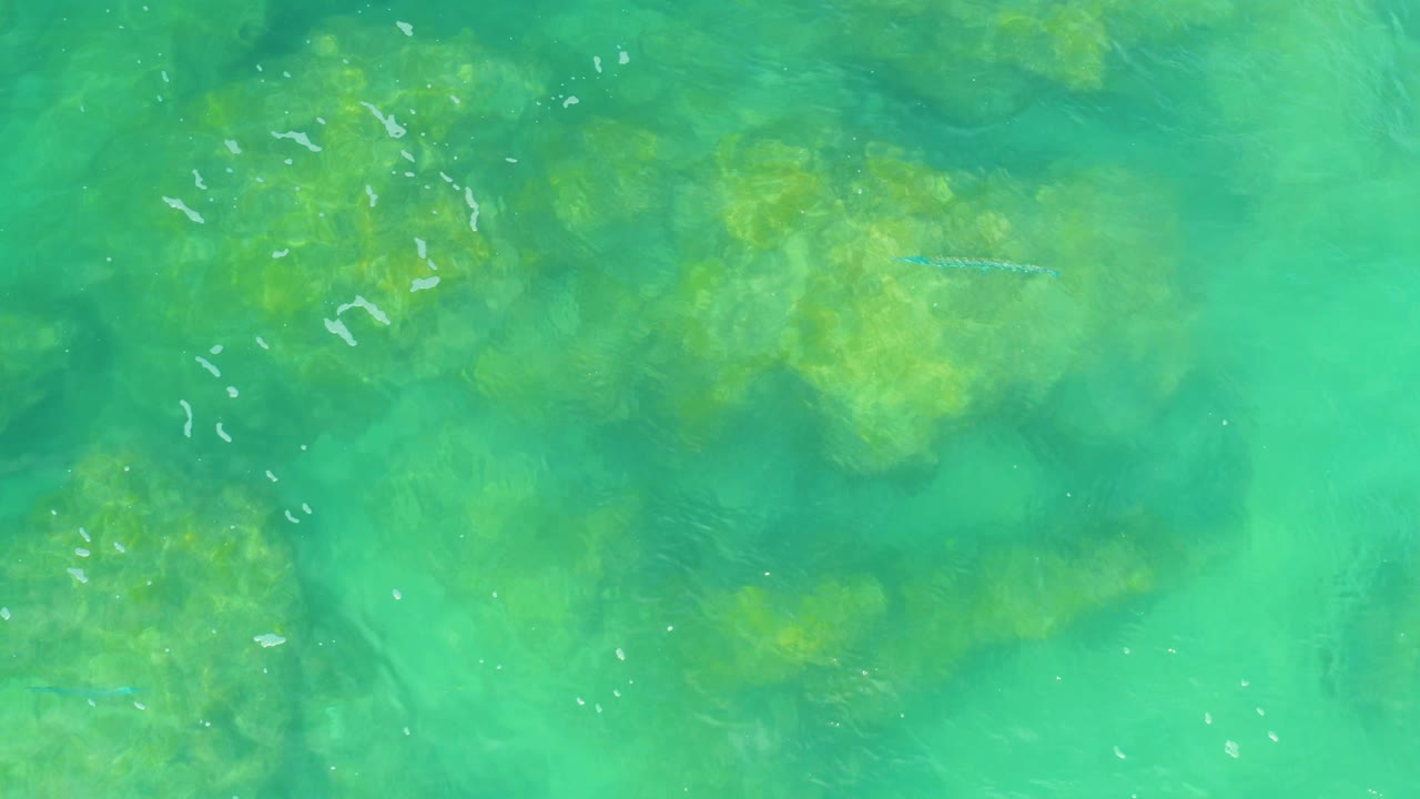Top-down Laguna Beach water frame showing blue-green surface color, darker submerged rock patches, faint wave streaks, and no visible people.