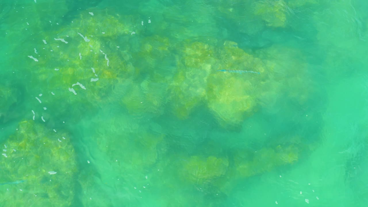 Overhead 4K frame from Laguna Beach, Baja California Sur, with reef-like rocks under clear green-blue water and scattered white ripples, no visible people.