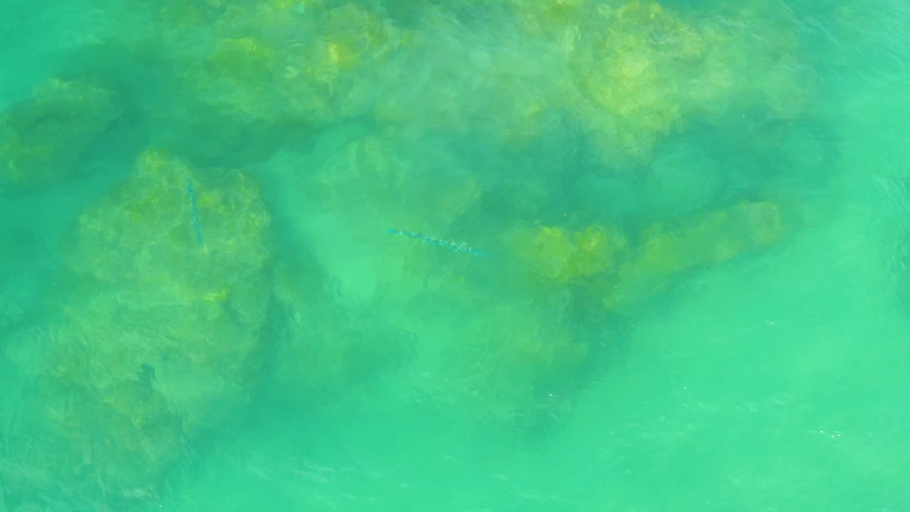 Aerial frame over green-blue Laguna Beach water, showing muted rock forms under the surface and faint pale specks in the water, with no visible people.