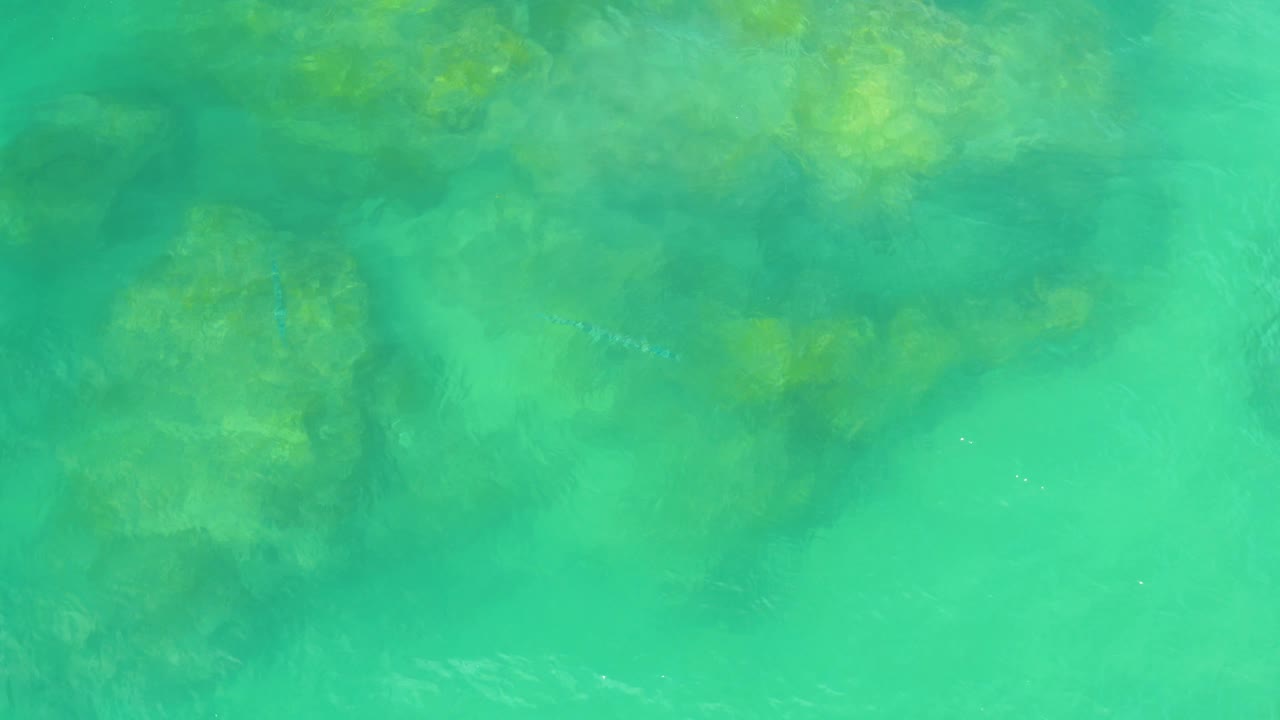 Top-down view of aqua coastal water in Laguna Beach, Baja California Sur, with darker underwater patches softened by ripples and no visible people.