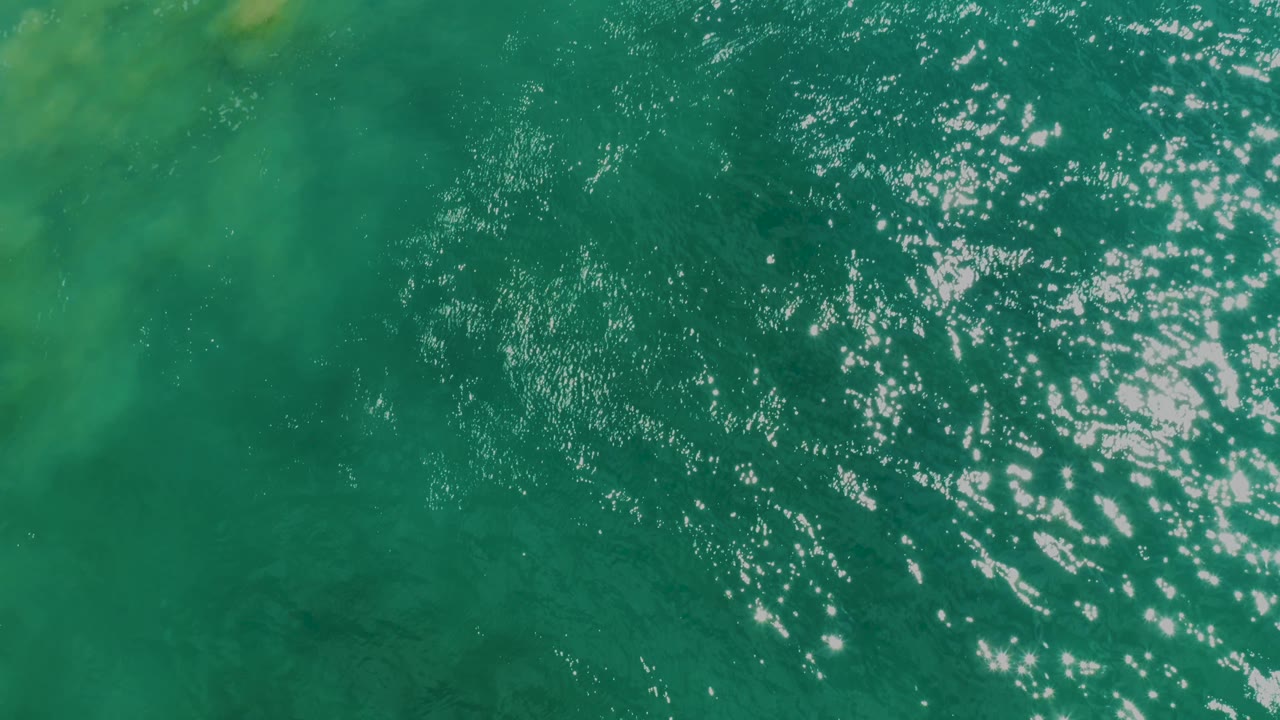 Top-down turquoise sea at Laguna Beach, Baja California Sur, shows mottled yellow-green sand or seaweed color beneath the ripples and no visible people.