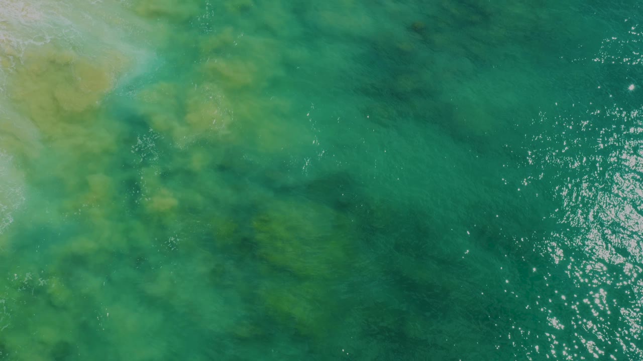 Top-down view at Laguna Beach with cloudy green and blue shallow water patterns, a small sandy corner near the upper left, reflected light on the right, and no visible people.