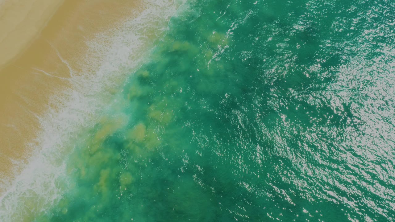 Overhead view of blue-green ocean water meeting pale sand at Laguna Beach, Baja California Sur, with white foam along the upper left shoreline and no visible people.