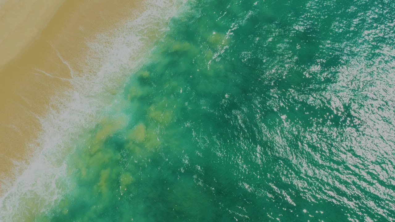 Aerial frame of a foamy wave edge sliding along pale sand at Laguna Beach, Baja California Sur, with textured teal water filling most of the view and no visible people.