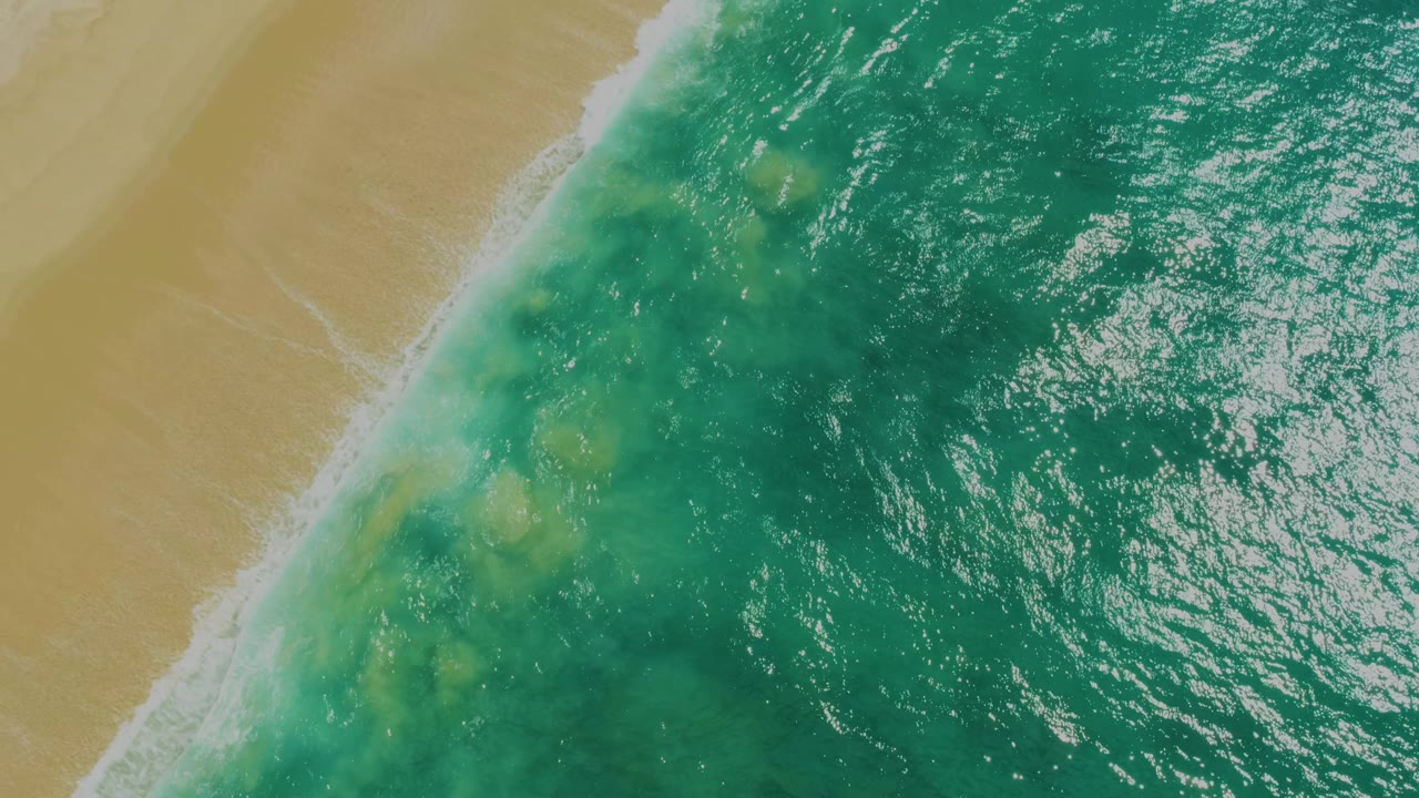 High overhead view of a diagonal white surf line beside rippled green-blue water and tan sand at Laguna Beach, Baja California Sur, with no visible people.
