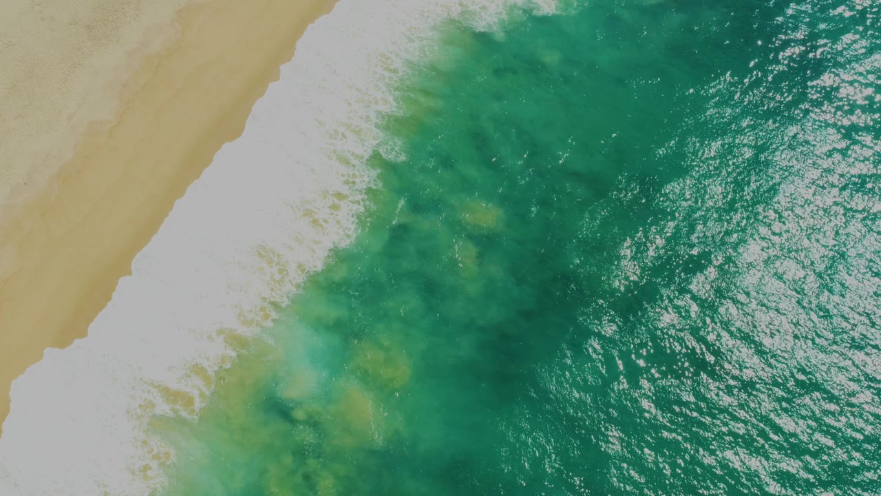 Aerial frame of green sea water spreading beside a diagonal band of white foam and tan sand at Laguna Beach, Baja California Sur, with no visible people.