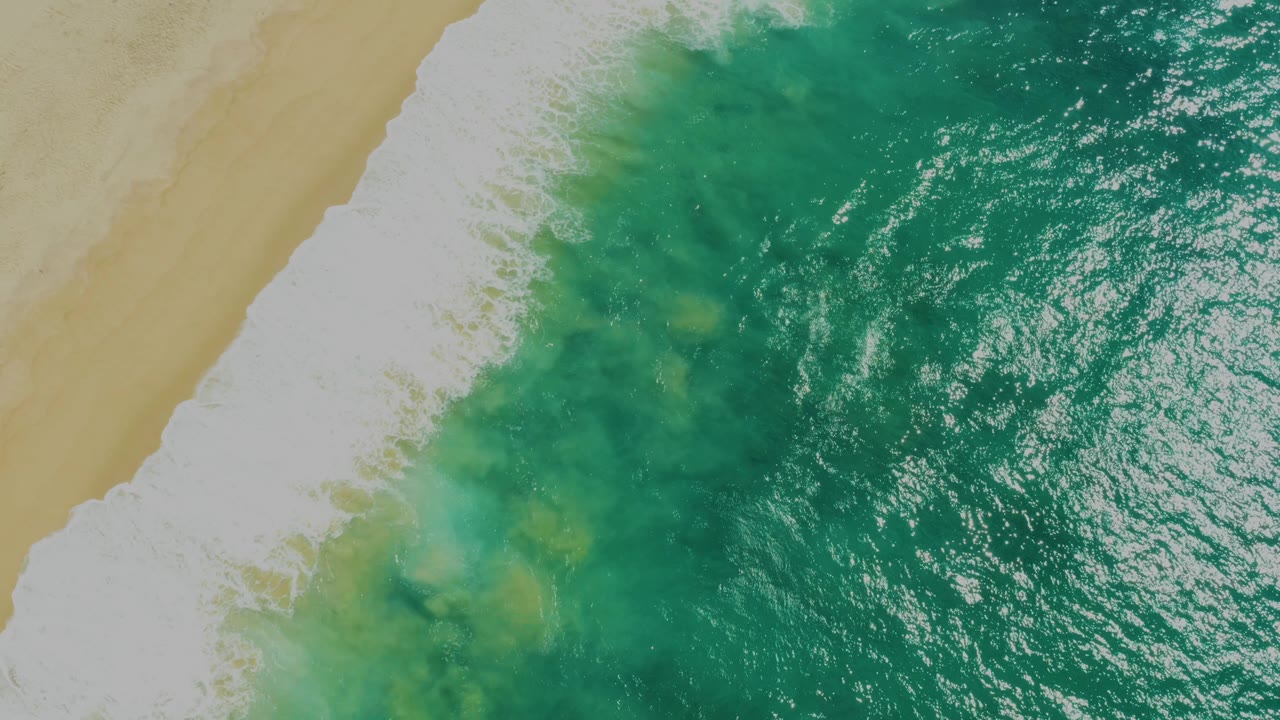 Top-down view of bright green sea water, white breaking surf, and a narrow tan beach edge at Laguna Beach, Baja California Sur, with no visible people.