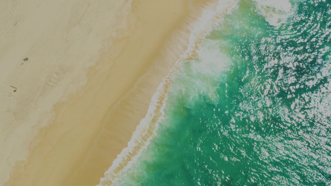 Aerial view of white shoreline foam beside green-blue rippled water and smooth tan sand in Laguna Beach, Baja California Sur, with no visible people.