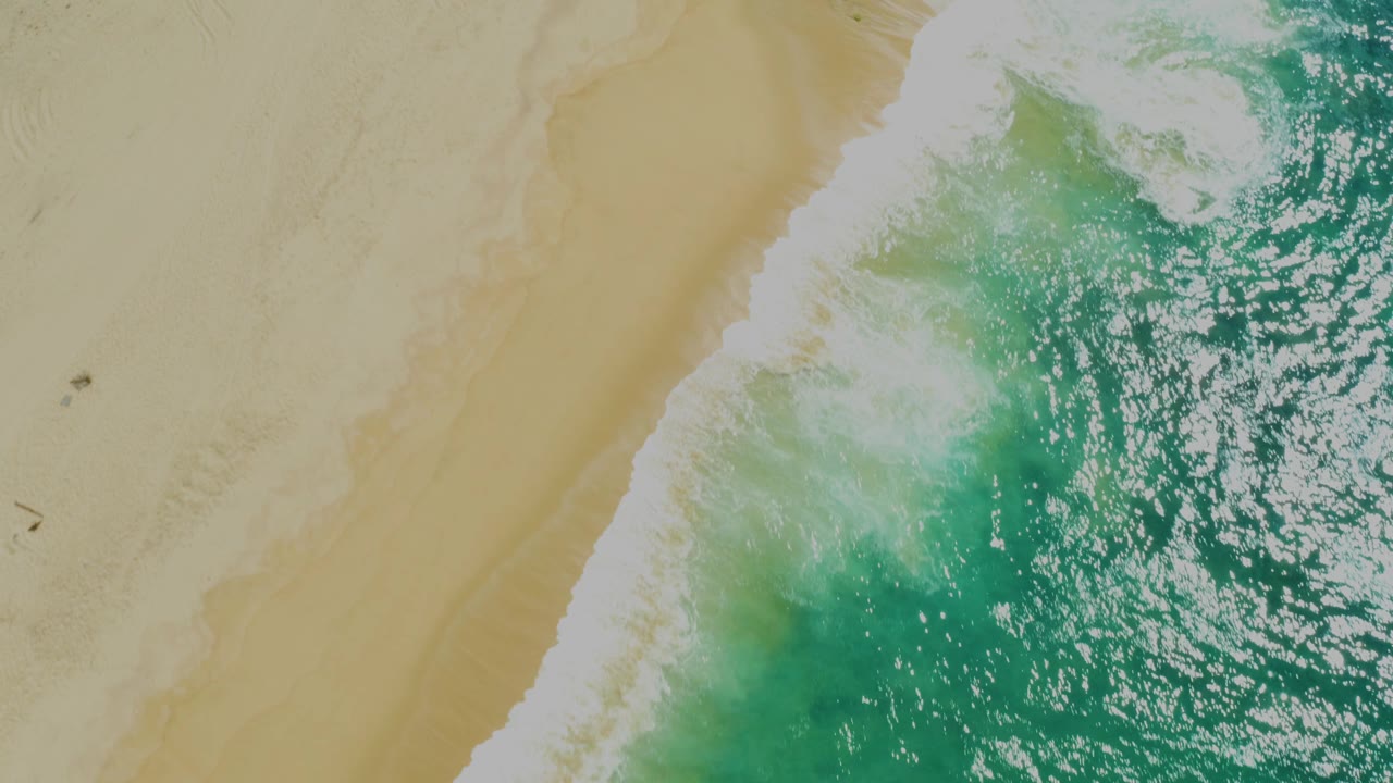Bright overhead shoreline frame with foamy surf, pale tan sand, and textured turquoise water at Laguna Beach, Baja California Sur, with no visible people.