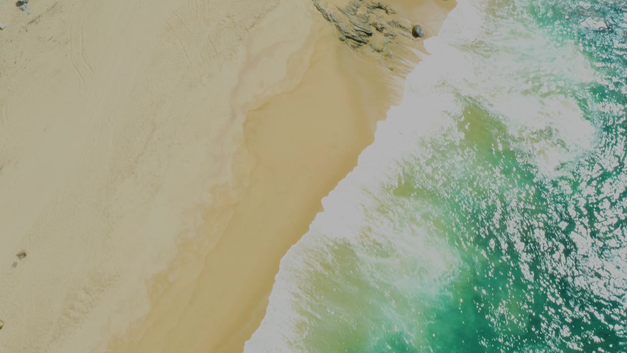 Top-down beach view where white foam crosses the sand beside green water at Laguna Beach, Baja California Sur, with no visible people.