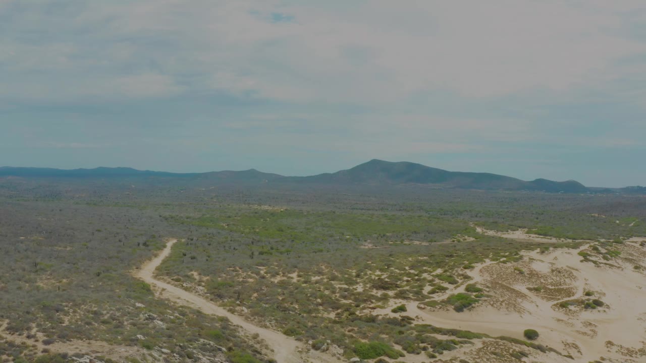 Aerial view in Laguna Beach, Baja California Sur with the sandy wash more prominent at lower right, a winding track at left, scattered scrub, distant mountains, and no visible people.