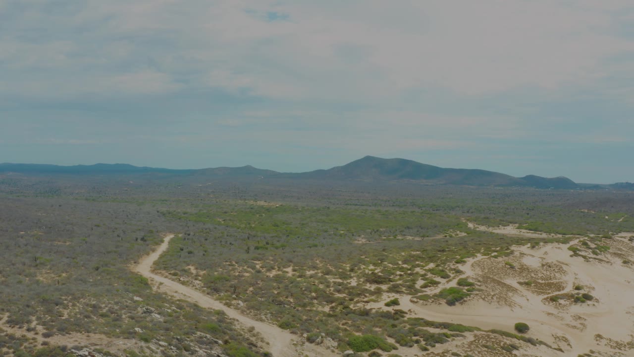 Wide aerial frame from Laguna Beach, Baja California Sur showing the sandy trail bending through scrubland, a broad flat midground, hazy mountain forms on the horizon, and no visible people.