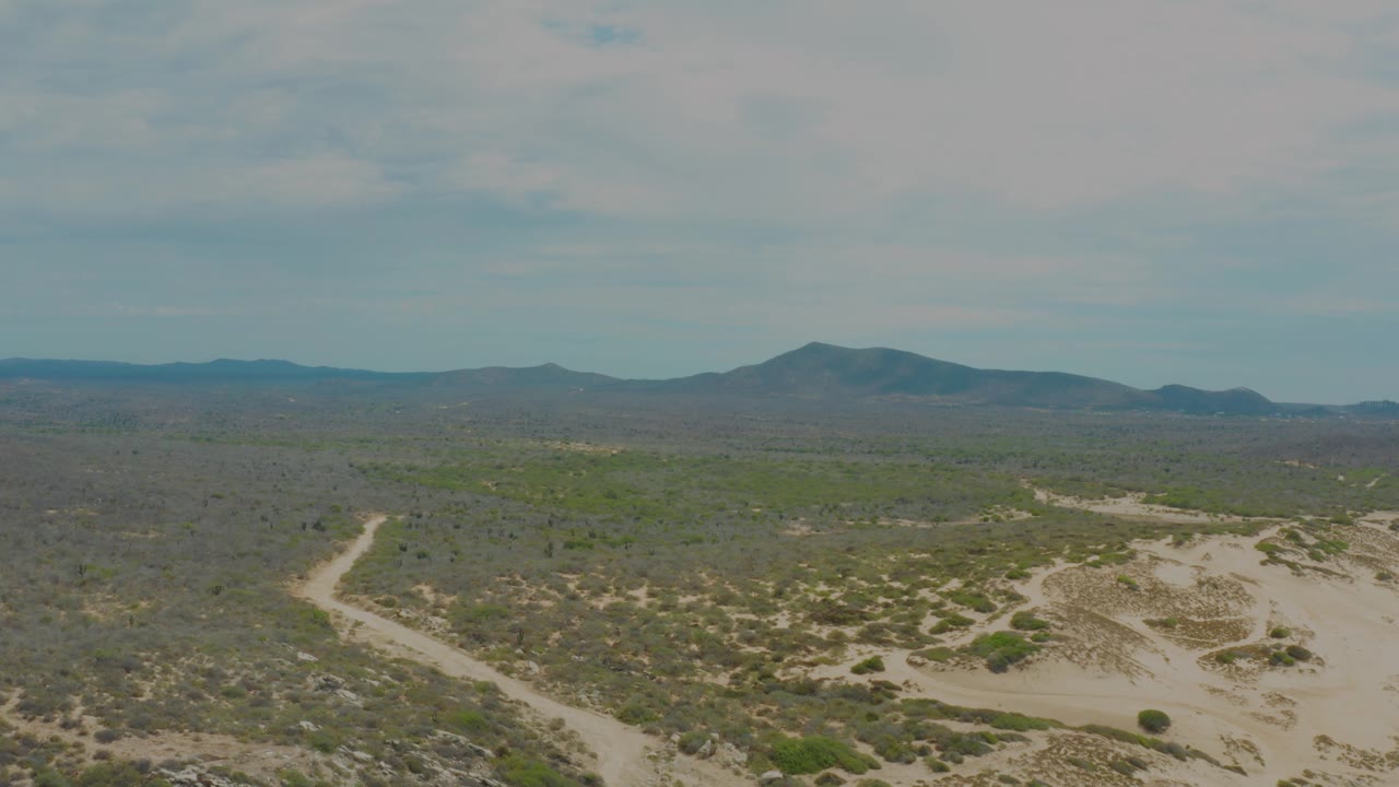Aerial frame over Laguna Beach, Baja California Sur with the sandy path visible at lower left, scrub-covered flats across the center, a dark mountain ridge beyond, and no visible people.