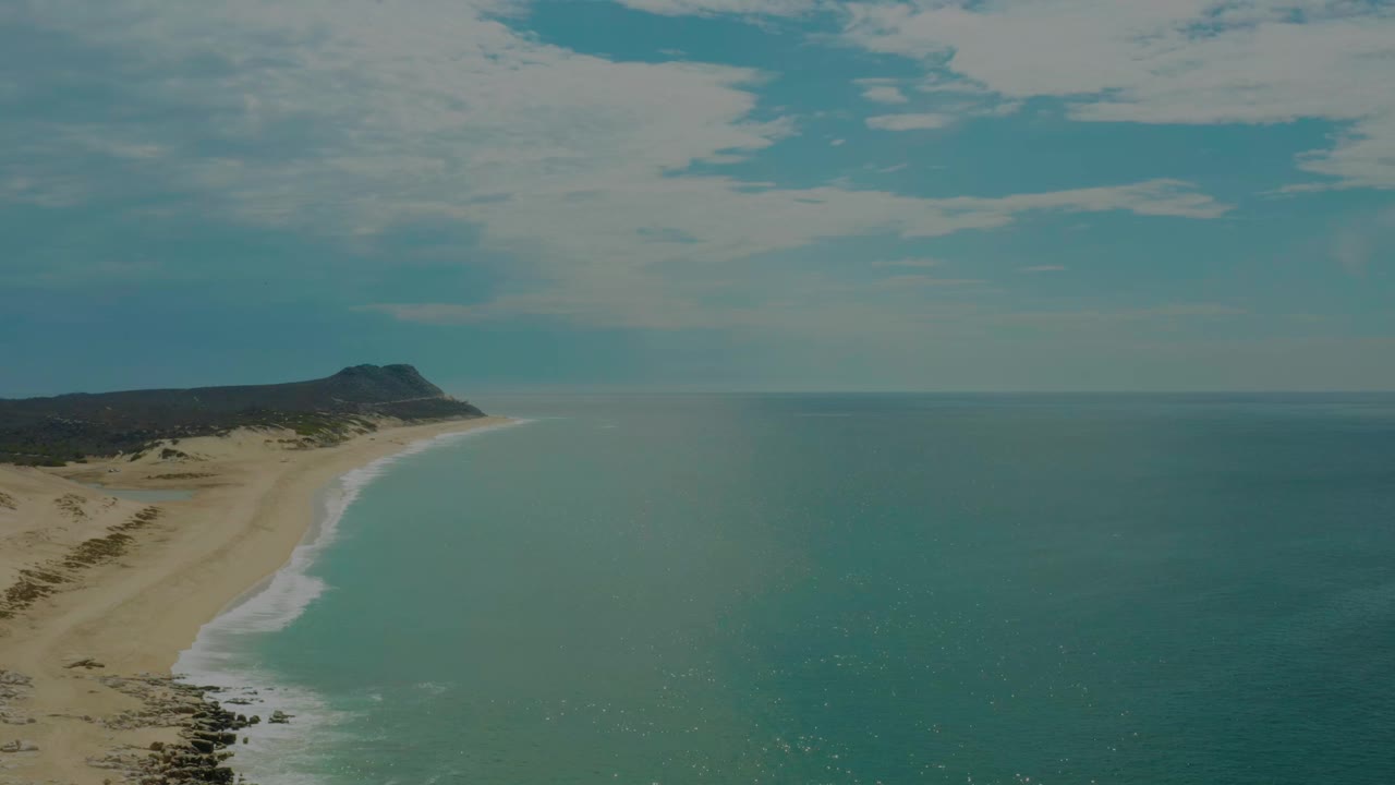 Opening aerial frame over Laguna Beach in Baja California Sur, with a sandy shoreline bending toward a distant low headland, blue-green water, clouded sky, and no visible people.
