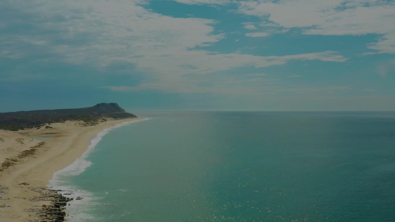 Aerial frame over Laguna Beach in Baja California Sur with mild waves reaching the sand, the distant headland still left of center, broad ocean to the right, and no visible people.