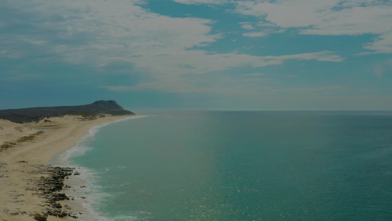 One second into the clip, the Laguna Beach coastline remains framed with tan sand at left, jagged dark rocks near the lower shore, blue-green water extending to the horizon, and no visible people.
