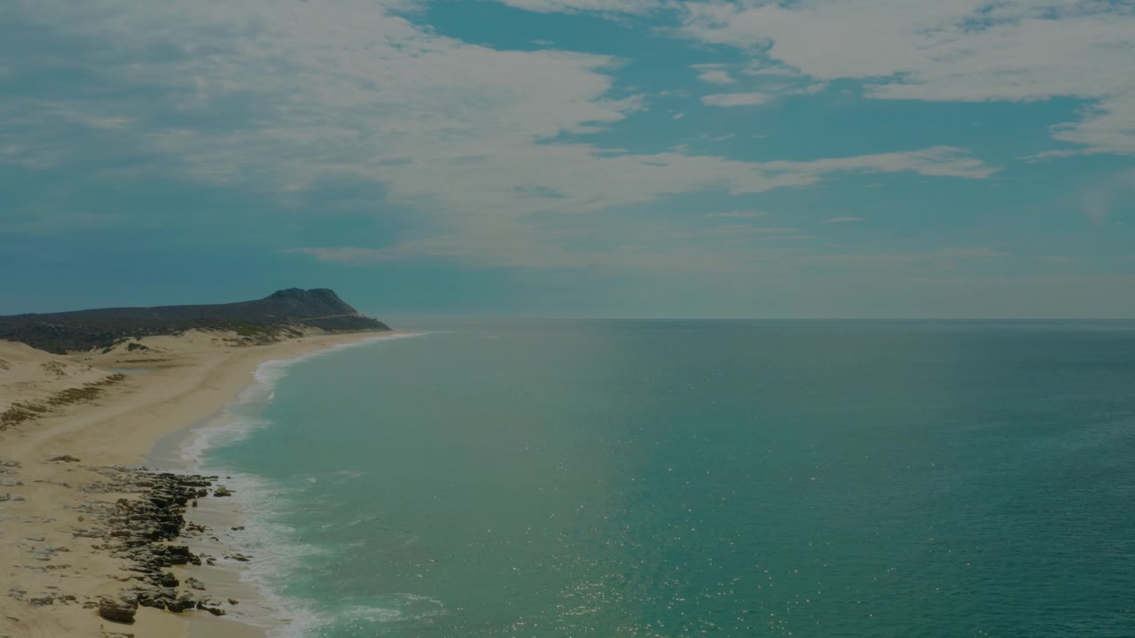 At 0:02, the aerial view shows the curving sandy shoreline of Laguna Beach, Baja California Sur, with a thin band of white surf, scattered rocks below, open sea to the right, and no visible people.