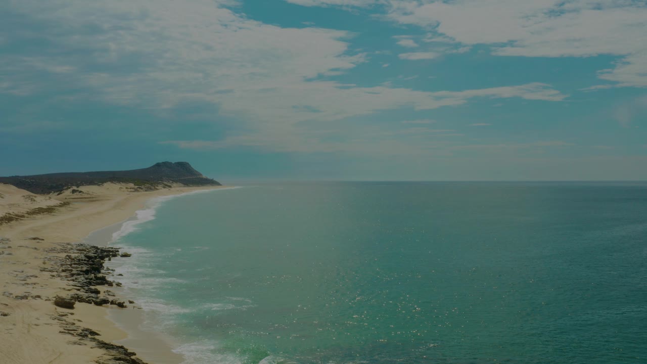 Opening frame of Laguna Beach, Baja California Sur, with sand and dark shore rocks on the left, blue-green ocean on the right, a distant headland, and no visible people.
