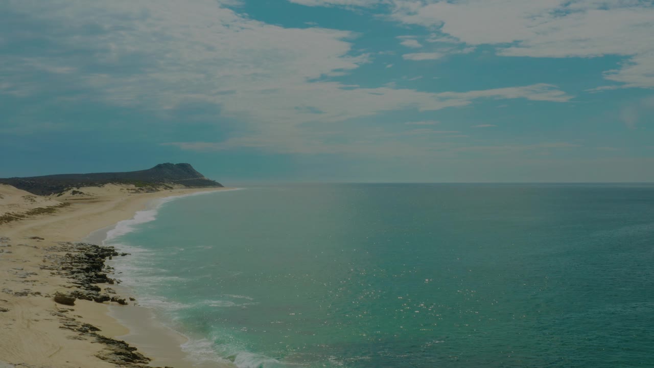 A bright aerial view over Laguna Beach, Baja California Sur, with ocean texture across the right side, sandy coast at left, a dark rocky strip near the water, and no visible people.