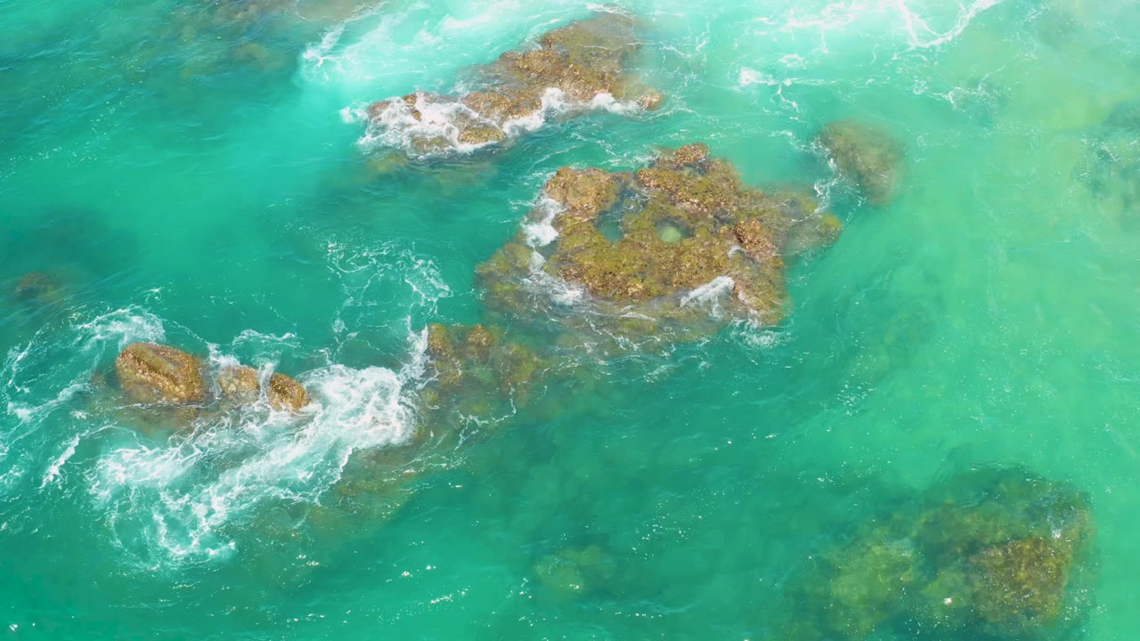 Aerial top view at Laguna Beach, Baja California Sur, showing blue-green water, clustered rocks near center, white surf curling from the lower left, and no visible people.