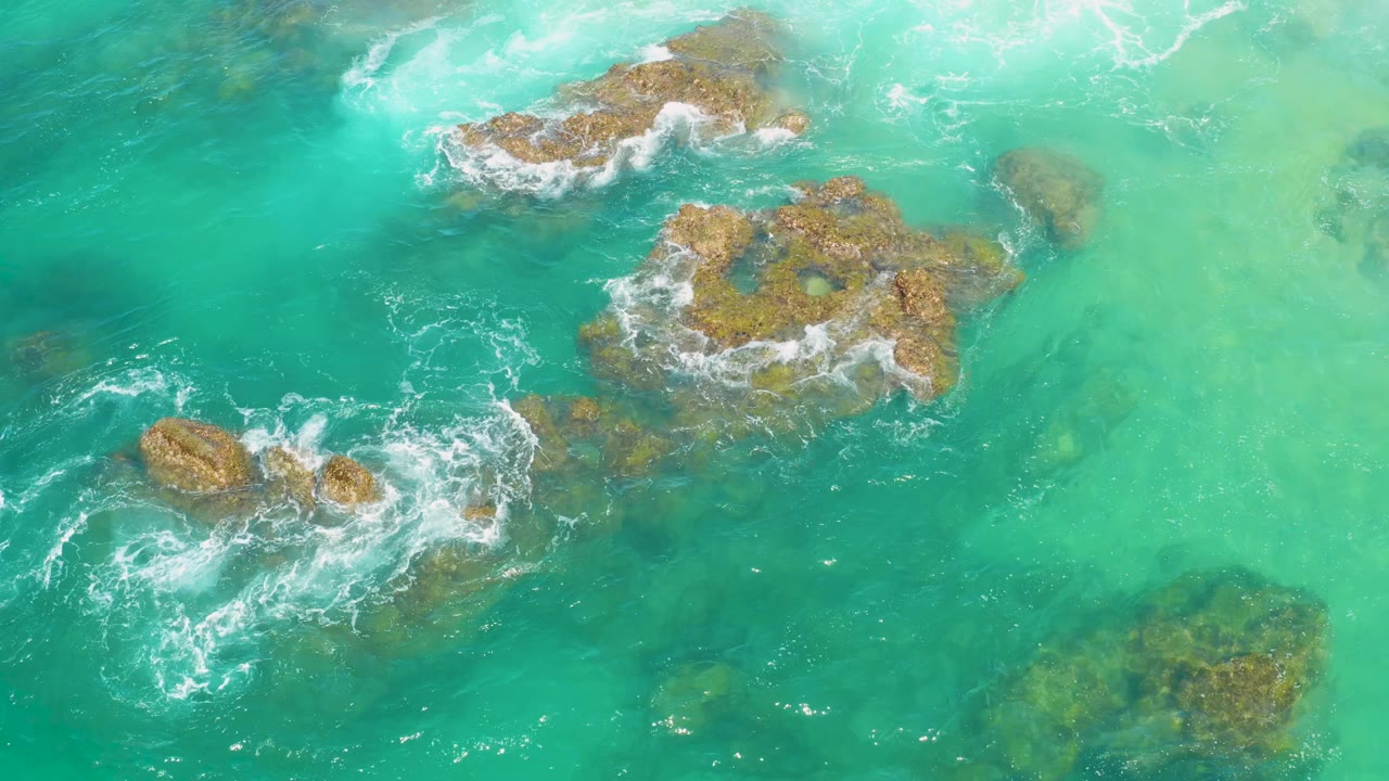 Top-down Laguna Beach, Baja California Sur water view with pale turquoise shallows on the right, darker blue water on the left, rocky outcrops, streaks of surf, and no visible people.
