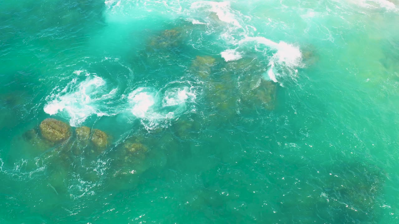 Bright green ocean water surrounds low brown rocks as white foam breaks across the upper and center areas at Laguna Beach, Baja California Sur, with no visible people.