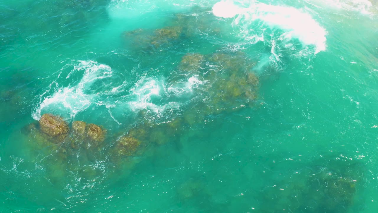 Several exposed and submerged rocks sit under green water while white wash trails diagonally through the overhead view at Laguna Beach, Baja California Sur, with no visible people.