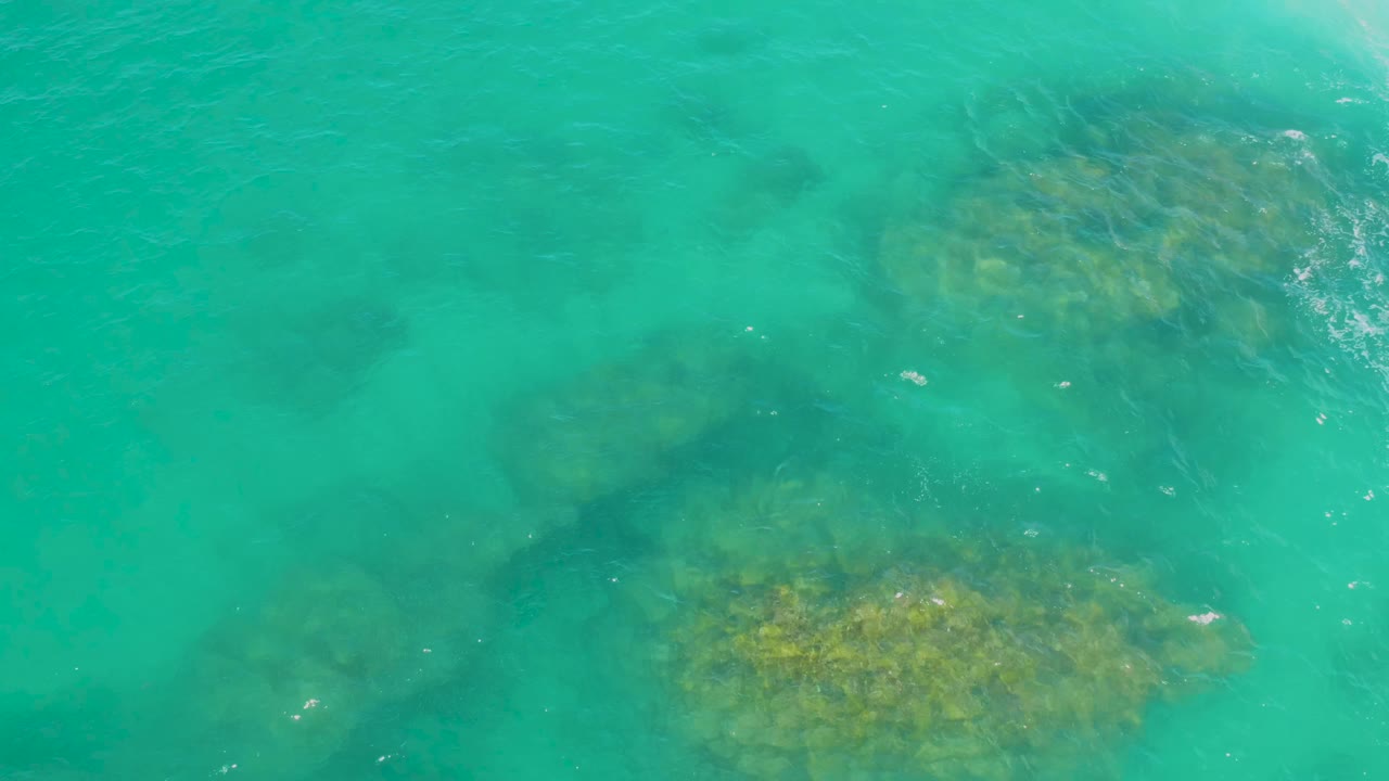 Clear green ocean water reveals darker rock shapes below the surface in Laguna Beach, Baja California Sur, with a yellow-brown formation lower in frame and no visible people.