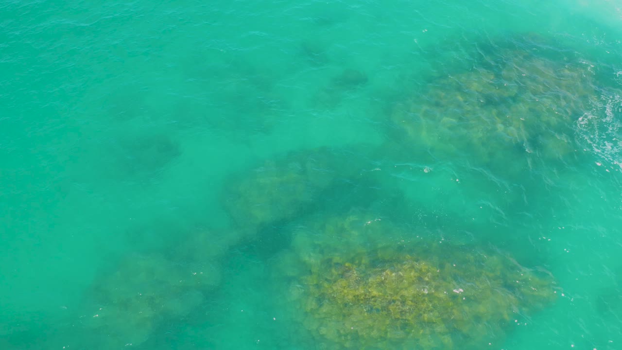 Turquoise-green water covers clustered submerged rocks at Laguna Beach, Baja California Sur, with small white ripples near the right side and no visible people.