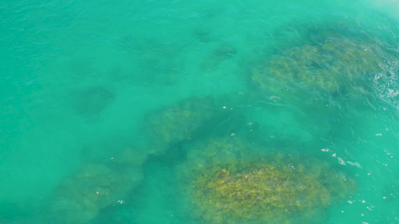 Overhead view of green water at Laguna Beach, Baja California Sur, with large submerged rocks spread across the frame and faint white wave texture on the surface, no visible people.