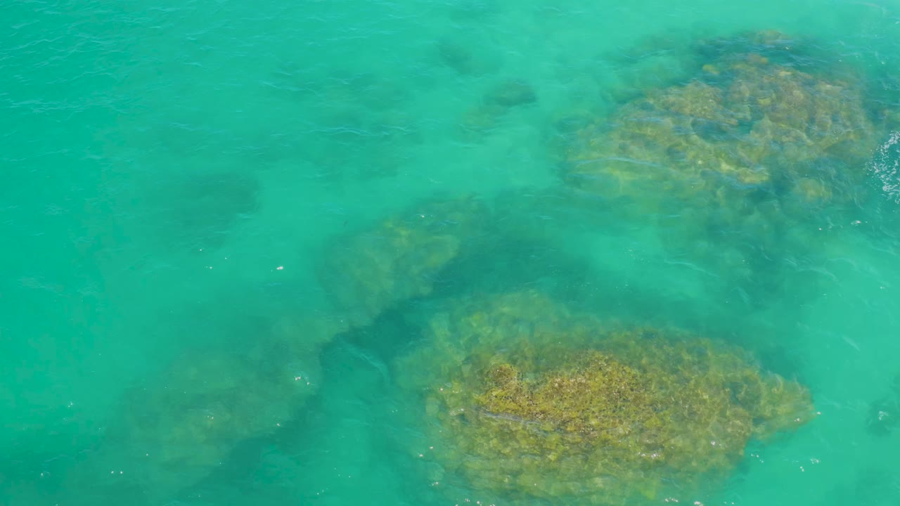 Aerial frame over clear turquoise water at Laguna Beach, Baja California Sur, where reef patches and darker underwater areas are visible, with no visible people.